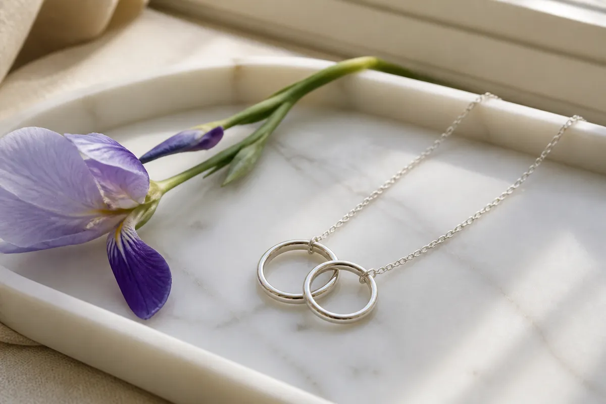 A sterling necklace with two interlocking circles laid on a white marble tray beside a sprig of iris, window light