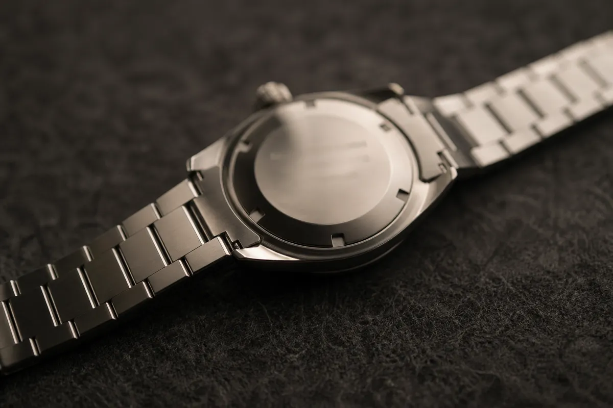 A steel automatic watch lying face down on dark felt, its small caseback engraving softly out of focus, moody light