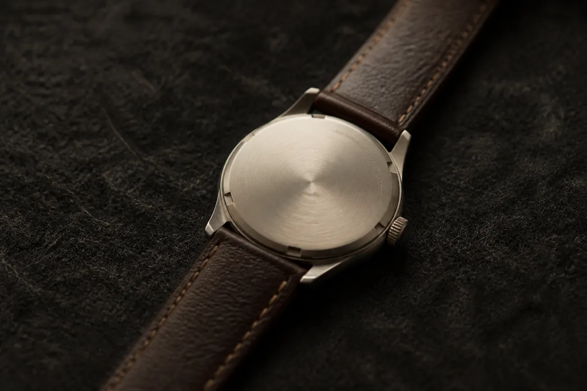 A leather-strap watch lying face down on dark felt, its small caseback engraving at an angle and softly out of focus, moody light