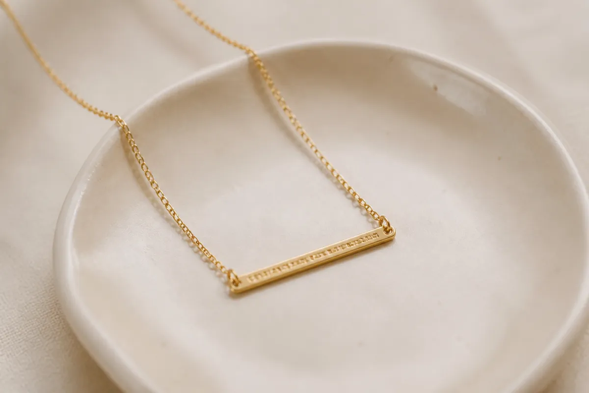 A dainty gold bar necklace with tiny stamped coordinates slightly out of focus, laid on a cream ceramic dish