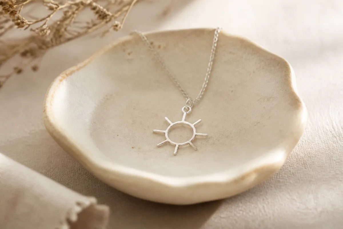 A dainty silver pendant shaped like a child's simple doodle of a sun, on a small ceramic dish
