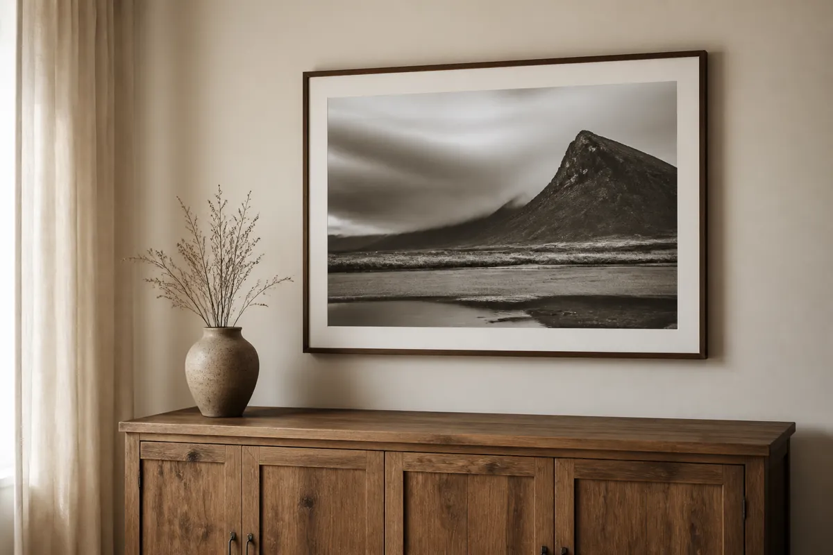 A large framed black and white landscape photograph hanging on a wall above a wooden sideboard with a vase